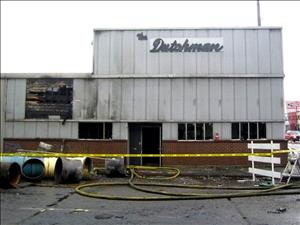 A front view of the fire-damaged The Dutchman rehearsal and recording studio. The sign on the second story front says The Dutchman. The entrance is blocked by yellow emergency tape, fire hoses snake along the pavement, and various pieces of unidentifiable wreckage litter the ground in front.